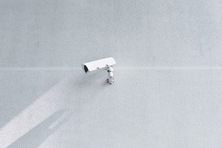 A photograph of a CCTV surveillance camera attached to a blank wall and its shadow