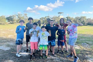 group of 8 campers and 2 activity leaders stand with their palms facing camera to show off their paint covered hands. all are looking at the camera and smiling. the rock climbing tower is seen in the background