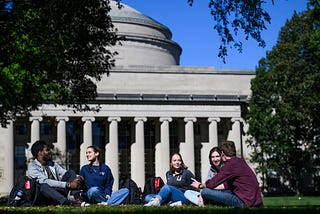 Five students sit on the grass with the MIT Dome behind them