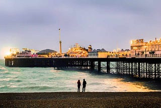 A man and a woman standing by the shore with a view of Brighton Pier, lit up for the evning