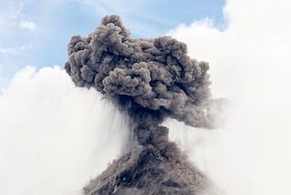 A gray cloud of smoke coming out of a volcano against a blue sky with white clouds.