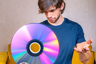 White man with shaggy hair and a bit of facial hair who is probably in his 20s is wearing an athletic blue t-shirt and holding a laser disc in one hand. His other hand is raised inquisitively and his stare is fixed on the laser disc with a facial expression that indicates he’s thinking something along the lines of, “What the fuck am supposed to do with this thing?” Image was AI generated.