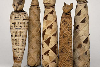 A row of five mummified cats in the British Museum. Each one is standing on a small square plinth, and essentially resembles a long tube wrapped in strips of (presumably) papyrus, in a variety of patterns, and with a carved (wooden?) cat’s head at the top
