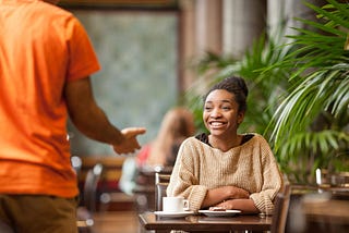 A student sitting in a cafe and smiling as a friend approaches.