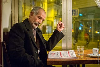An older man sits in a cafe reading a newspaper.