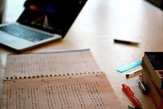 A laptop open next to a notepad with pages filled in handwritten notes.