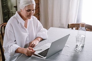 An elderly lady with white hair is sitting at a table and looking at her laptop. In her hand she holds a credit card.