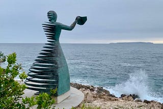 A bronze metal sculpture of a woman holding a handkerchief out as she faces the sea. On one side of the sculpture the metal is cut into horizontal ribbons. A wave breaks below on the rocky shore.
