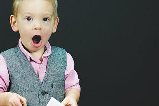 A well-combed little blonde boy — who is wearing a gray vest and pink seersucker collared shirt — looks up from the book he is holding and gapes his mouth open in amazement.