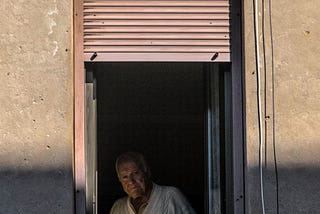 old man sitting in darkness, wearing a white shirt, looking out of his window.