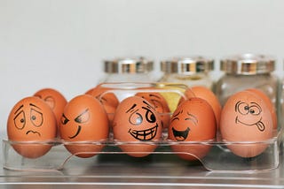 Eggs with faces of different emotions drawn on them, sitting in a transparent holder.