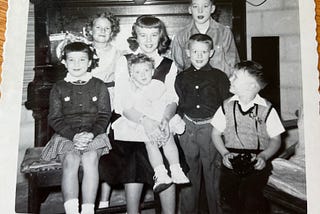 Seven children in the 1950’s pose for a photo by a piano.