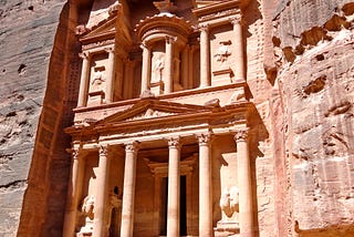 A pink-hued building in the Roman-Greek style with Corinthian columns is carved into a flat rockface
