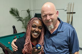 Partners Amani and Doug pose for a picture in front of a pool table while holding the 2 ball