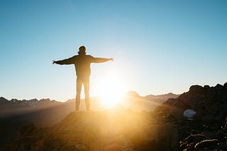 Silhouetted person standing on rock with arms outstretched, the sun rising in front of them.