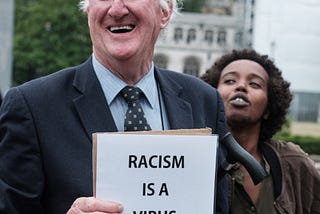 White man holding a sign smiling and a black woman standing beside him. The sign reads “Racism is a virus, we are the vaccine.”