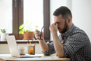 Photo of a stressed out man, sitting at a desk, in front of a laptop. He has his glasses in his right hand and, with his left, is squeezing the bridge of his nose. Photo courtesy of Freepik