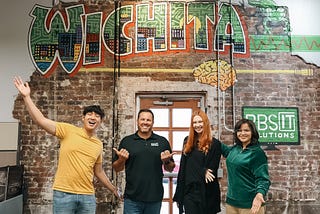 Three young people and one IT professional pose joyfully in front of a mural that says “Wichita.”