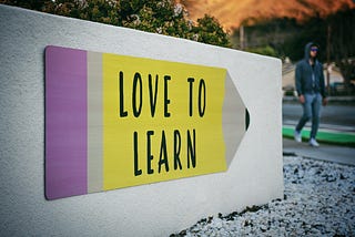 Picture of a white small wall with a sign on it in the shape of a pencil. On the sign it reads “love to learn”.