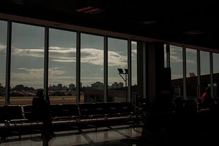 Foto em um aeroporto. Na esquerda, a silhueta de uma moça arrumando o cabelo; na direita, um homem vestido de vermelho.