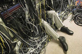 A person has their whole body immersed under a pile of random cables connecting systems in a random way. There are hundreds of cables and the person’s legs are sticking out, they are wearing beige trousers and black shoes.