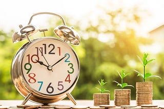 Image of a colorful analog alarm clock and three stacks of coins increasing in size with plant sprouts on them.