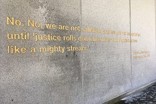 An inscription on a wall in San Francisco’s Dr. Martin Luther King, Jr. memorial reading, “No. No, we are not satisfied, and we will not be satisfied until ‘justice rolls down like water and righteousness like a mighty stream.’ Washington, D.C., 1963”.