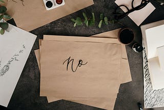A wooden desk with a coffee cup and a sheet of brown paper with cursive handwriting that says “no.”