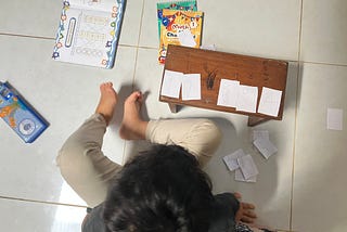Seorang anak kecil duduk di lantai sedang belajar matematika dengan menggunakan potongan kertas bertuliskan angka. Di depannya terdapat meja kecil kayu dengan angka 1, 2, 6, 9, dan 4 yang ditempel berurutan. Di lantai juga terlihat buku latihan matematika anak, beberapa kertas angka lainnya, dan alat tulis. Suasana menunjukkan aktivitas belajar sambil bermain di rumah