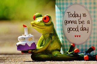 Ceramic frog holding a birthday cake in front of a mug reading “Today is gonna be a good day”