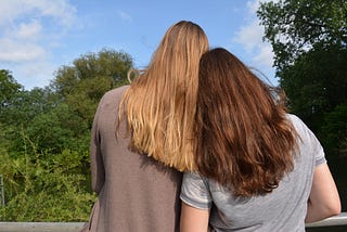Good friends enjoying time outdoors. Photo shows two women, one blonde and one brunette, with their backs to the viewer, looking out to a place with vegetation