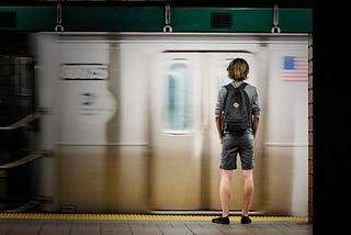 A young person with a backpack stands with back turned to the viewer on a subway platform.
