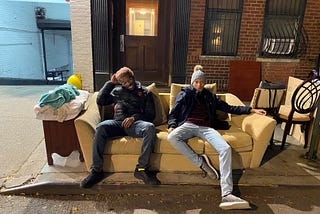 author (on left) and his friend (on right) sitting on a beige couch that is on a sidewalk. There is a chair and a cabinet on the sidewalk next to the couch