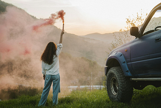 Woman holding a red smoke flare stands beside an off-road vehicle on a grassy hill against a mountain backdrop at sunset