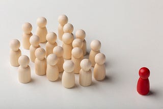 A cluster of pale wooden pegs on a grey background. An identical wooden peg but painted red stands alone, some distance away.