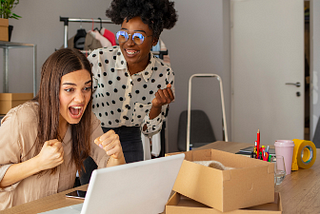 Two women are at a wooden table in front of a laptop excited at what they’re looking at on the laptop. They’re in an office with a fashion rack behind them.