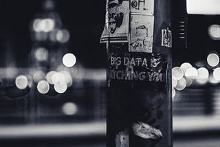 An ominous black and white image of a lamppost with posters and stickers plastered across it, including the phrase ‘BIG DATA IS WATCHING YOU’