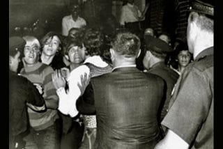 black-and-white photo of a crowd of people pushing with their hands. someone wearing a cop’s hat is facing a group of young people who are being pushed.