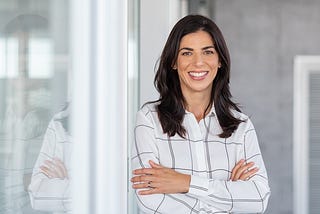 A professional woman smiling confidently while standing with her arms crossed. She has long dark hair and is wearing a white blouse with a black grid pattern. The background shows a modern office space with glass walls, providing a clean and contemporary setting.