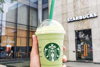 A hand holds up a green, frothy Starbucks beverage in front of an industrial-looking Starbucks. Maybe they’re about to Pay It Forward?