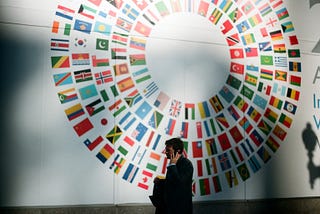 A man speaking on his cellphone walking in front of national flags of countries arranged in circular shape.
