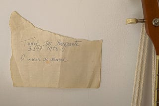 Na parede, um pedaço de papel rasgado e amarelado diz “Tunel da Imigrante. 3.147 mts. O mair do Brasil”. Ao lado, pode-se ver parte do braço de um ukulele pendurado na mesma parede por cordas de algodão cru.