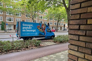 This is a picture of a blue delivery truck with a picture of fresh food on it and Dutch words that say, “Geoogst, geplukt en bezorgd,” which means, “harvested, picked and delivered.”