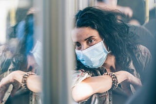 A woman in a surgical mask looks pensively out the window of a bus.