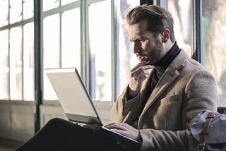 A white man in his 30s wearing a brown jacket and black turtleneck stares at his laptop as though pondering a dilemma.