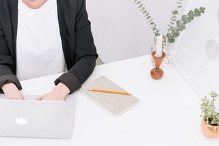 Person sitting at bright desk with candle and green plants while typing on laptop next to notebook and pencil