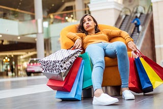 A woman sitting in a chair in the middle of a shopping centre, her hands full of bags of shopping and a look of exhaustion on her face.