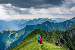 Two people walking a green mountain range with sunlit clouds cascading overhead.