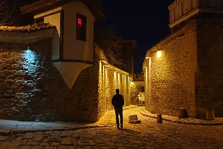 A man walks down a cobblestone road at night, buildings are on both sides of the narrow roadway.