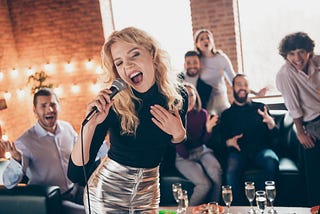 Pretty blonde woman in shiny shirt sings Karaoke infront of a crowd of friends.
